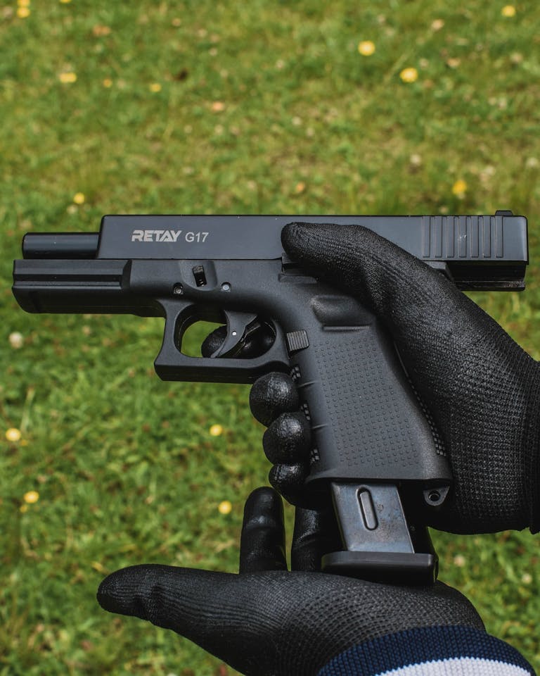 Close-up of a gloved person holding a firearm outdoors on grass.